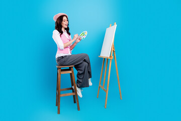 Fototapeta premium Young female artist painting on a blank canvas at an easel with palette and brush, wearing a pink beret and vest while sitting on a stool in a bright blue studio