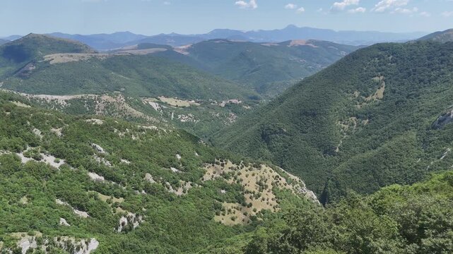 Paesaggio montano con vegetazione verde e fitta, colline sovrapposte e vallate naturali sotto un cielo azzurro con poche nuvole. Scena ampia, luminosa e incontaminata