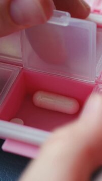 Close up person hand opening small compartment of pink plastic pill organizer containing capsule, showing daily management of medication, supplements for health. Person opening pink pill organizer box