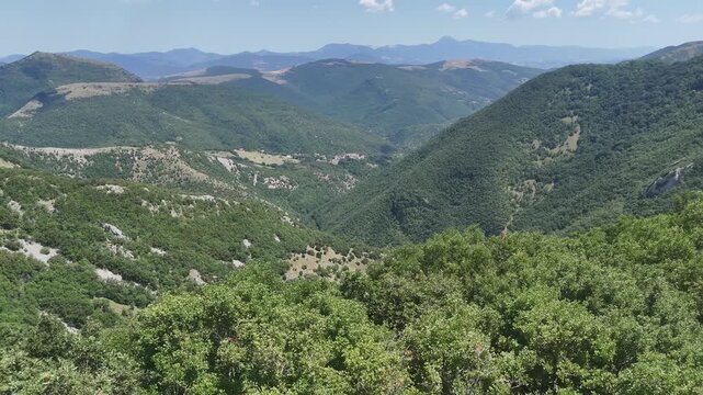 Paesaggio montano con vegetazione verde e fitta, colline sovrapposte e vallate naturali sotto un cielo azzurro con poche nuvole. Scena ampia, luminosa e incontaminata