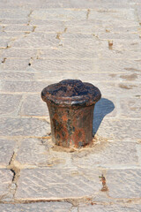 Fototapeta premium A vertical shot of an old rusty black iron mooring bollard mounted on a traditional stone paved street or dock surface under sunlight