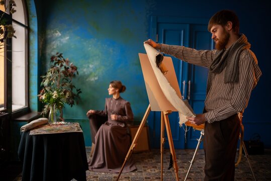 Male artist painting portrait of seated woman in vintage studio with teal wall and window light