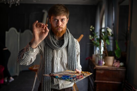 Bearded artist mixing oil paint on wooden palette in dim studio with plants and vintage furniture