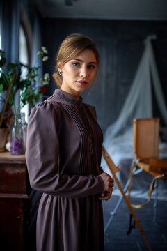 Young woman in vintage long dress standing in moody interior with studio props and natural window