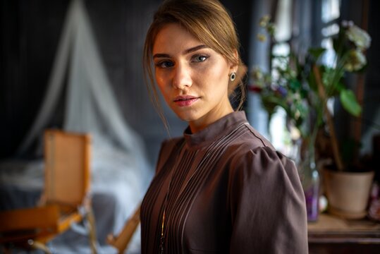An young woman in vintage long dress standing in moody interior with studio props and natural window