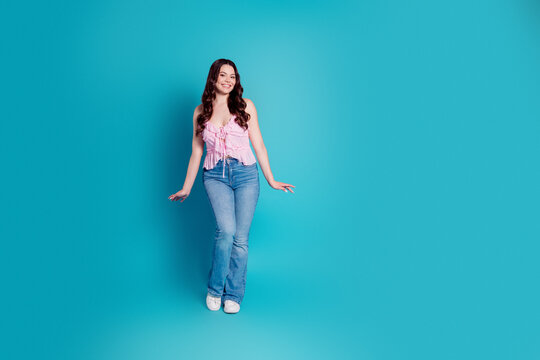 Smiling young woman in pink top and jeans posing against a turquoise studio background for casual fashion portrait conveying confidence and playful style