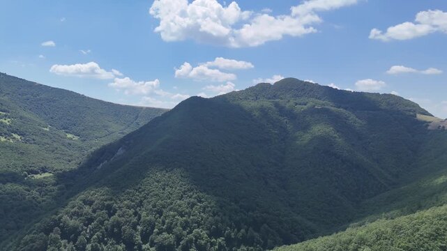 Montagne ricoperte di vegetazione verde con pendii morbidi, giochi di luce e ombra e cielo azzurro con nuvole bianche sparse