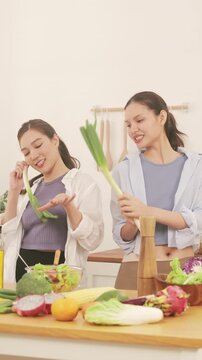 Happy young lesbian couple having fun and dancing with leeks while preparing healthy food in the kitchen.
