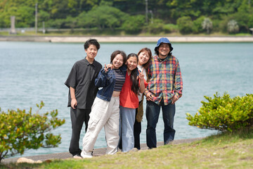 Five friends smile by a calm lake. They stand close, showing warm, joyful expressions. Green hills and shrubs frame the water's edge. Perfect for travel, family, or group outing themes © Masakazu Tokashiki