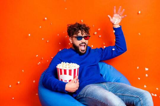 Excited man wearing 3D glasses eating popcorn on a blue bean bag against an orange background while watching a movie
