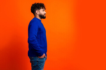 Young man in blue sweater standing in profile against bright orange background conveying casual confidence and thoughtful portrait mood © deagreez