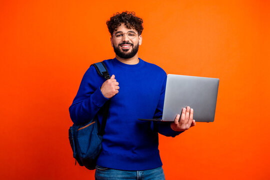 Young student holding laptop and backpack smiling in blue sweater against bright orange background, technology and education portrait conveying confidence and casual modern lifestyle