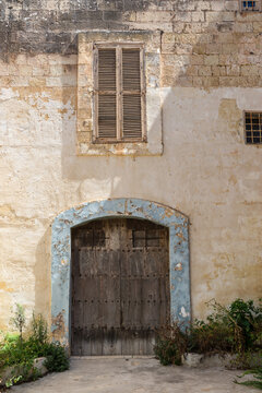 Historic limestone building with a weathered wooden door and shuttered window in the walled city of Mdina, Malta. Architectural details reflect the timeless charm and heritage of Malta's capital