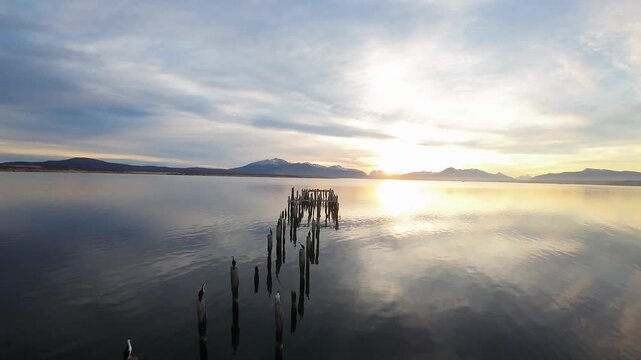 Historic Muelle Braun y Blanchard pier ruins rising from calm reflective water at sunset with snow-capped mountains and dramatic sky, Puerto Natales Chile, FPV aerial