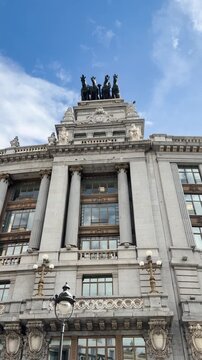 Upward view of historic stone building with columns and rooftop statues under blue sky
