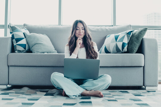 Young woman working on laptop at home sitting on floor in front of sofa, relaxed remote worker using computer in bright modern living room