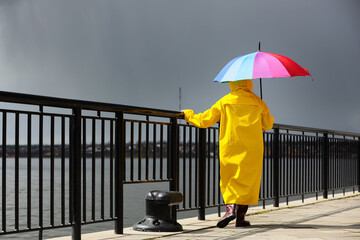 Obraz premium Woman in raincoat with rainbow umbrella near river on rainy day, back view