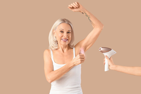 Mature woman with feather and hand holding photoepilator on beige background