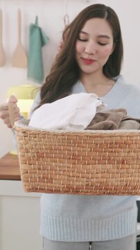 Happy young Asian woman holding a basket of folded clean clothes in a cozy home laundry room.