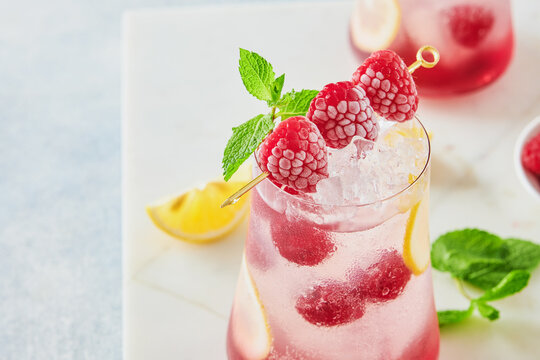 Close up of refreshing raspberry iced drink with lemon slices, mint leaves, ice cubes and fresh berries in elegant glasses on light marble table, bright summer beverage concept for cafe menu