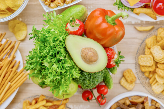 Flat lay of healthy vegetables with avocado, lettuce, broccoli and bell pepper next to burger, fries and ketchup, visual comparison of balanced nutrition and unhealthy fast food choices.