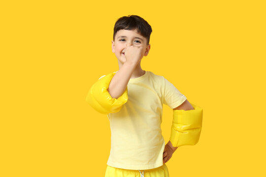 Cute little boy in inflatable armbands covering nose on yellow background