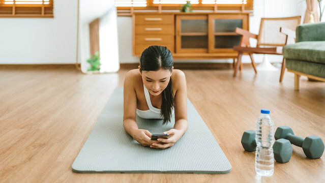 Young Asian woman lying on floor on yoga mat using smartphone at home. Beautiful smiling woman doing exercise relaxing while chatting with trainer online after workout