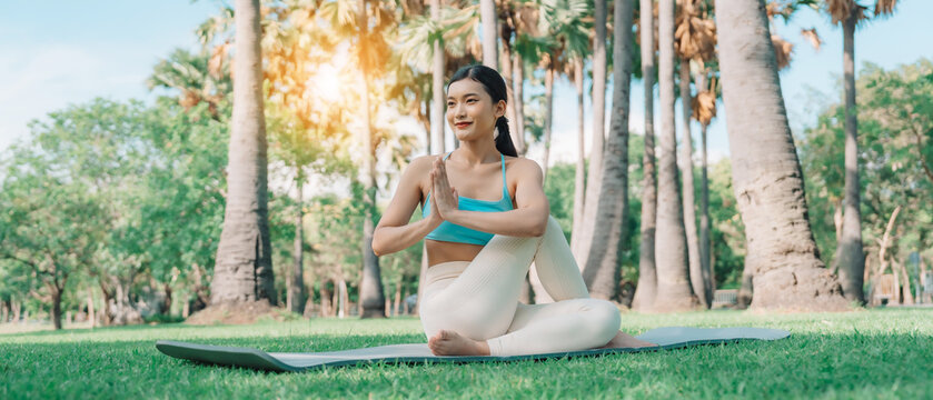 Asian woman meditating in a peaceful park setting, sitting cross-legged on green grass with trees in the background on a sunny day. Concept of mindfulness, self-care, and outdoor relaxation