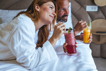 Relaxed Couple Enjoying Fresh Smoothie Breakfast in Hotel Bed Wearing White Bathrobes