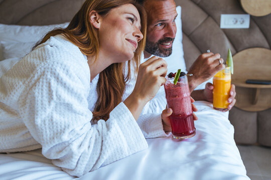 Relaxed Couple Enjoying Fresh Smoothie Breakfast in Hotel Bed Wearing White Bathrobes
