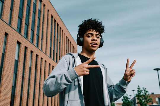 Happy teenage boy with curly hair wears headphones, grey hoodie outside. Young student listens to music, makes peace sign gesture with hands near modern brick building in city. Cheerful adolescent