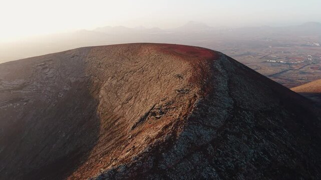 Close aerial view of the rim and inner slope of a volcanic crater at sunrise in Fuerteventura. Rocky volcanic terrain with the island landscape visible in the background, Canary Islands, Spain.