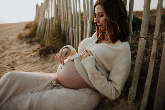 Expectant mother sitting on beach sand touching bare baby bump