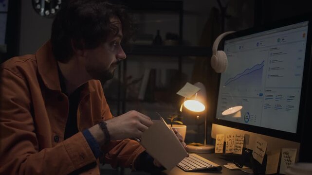 Caucasian young adult male office worker eating takeout food from box while working late at computer desk. Clip represents overtime work, late night shifts, and food delivery services