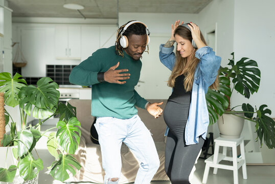 Man and pregnant woman wearing headphones dancing together in living room