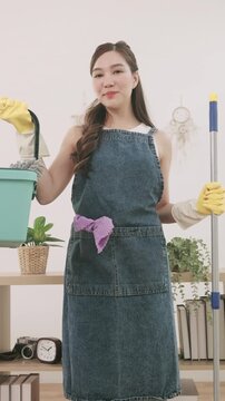 Smiling Asian woman ready to clean the house. She is holding a bucket and a mop, wearing an apron and rubber gloves.