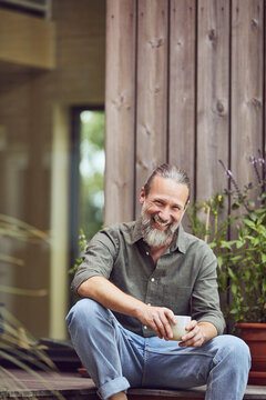 Cheerful mature man holding coffee cup while sitting outside tiny house