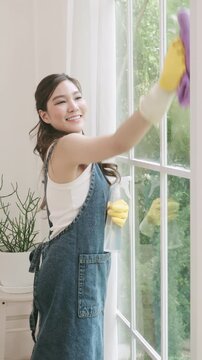 Happy Asian woman cleaning a window with a spray bottle and a microfiber cloth.