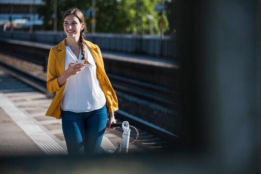 Smiling woman waits at train station with smartphone and scooter