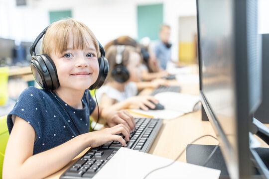 Smiling girl with bangs wearing wired headphones sitting in computer class at school