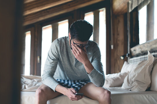 Tired young man with smartphone sitting on bed at home squinting