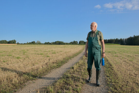 An elderly farmer wearing green dungarees walks along a dirt path in the morning, holding a water bottle and looking over his dried fields during a summer drought.