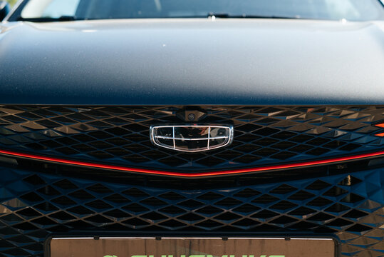 The front grille of a Geely car is prominently displayed along with its logo in a city environment. This image highlights the design details of the vehicle under bright sunlight Smolensk, Russia 07.17