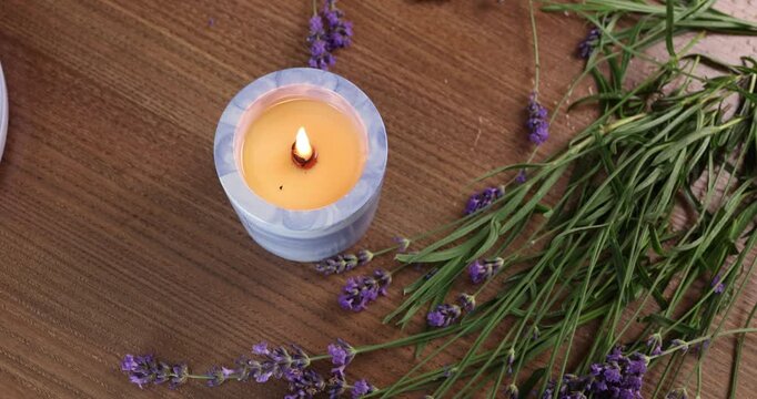 Burning candle flame lavender background video. Scented wax candle glows on wooden table with fresh purple blossoms. Cozy aromatherapy setup for spa relaxation or wellness mindfulness video footage.