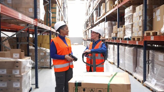 Two diverse female employees in safety vests and hard hats having a conversation in a large logistics warehouse among racks of packages
