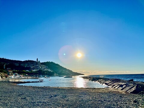 Playa de piedras frente a un pueblo costero italiano con el sol reflejado en el mar