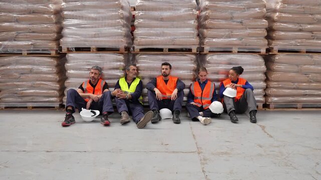 Diverse team of tired industrial workers sitting on the floor, resting and talking during a work break in a large logistics warehouse