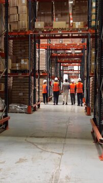 Group of logistics workers with hard hats and safety vests walking along a tall shelving aisle inside a large industrial warehouse