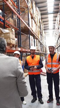 Manager leading a team meeting with warehouse workers. Staff in hard hats and safety vests listening to instructions in a storage aisle