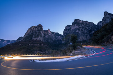 Obraz premium A long-exposure shot captures light trails from vehicles on a winding mountain road at twilight, surrounded by rocky peaks.